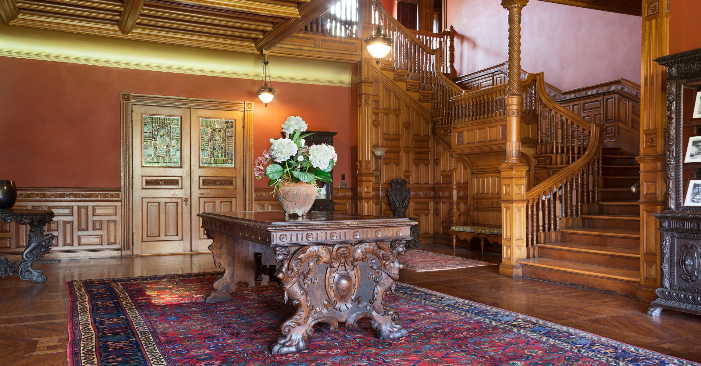 Wood paneled room with large staircase, stained glass panels in the doors, and a dark wood, heavily carved table on a red floral patterned carpet.