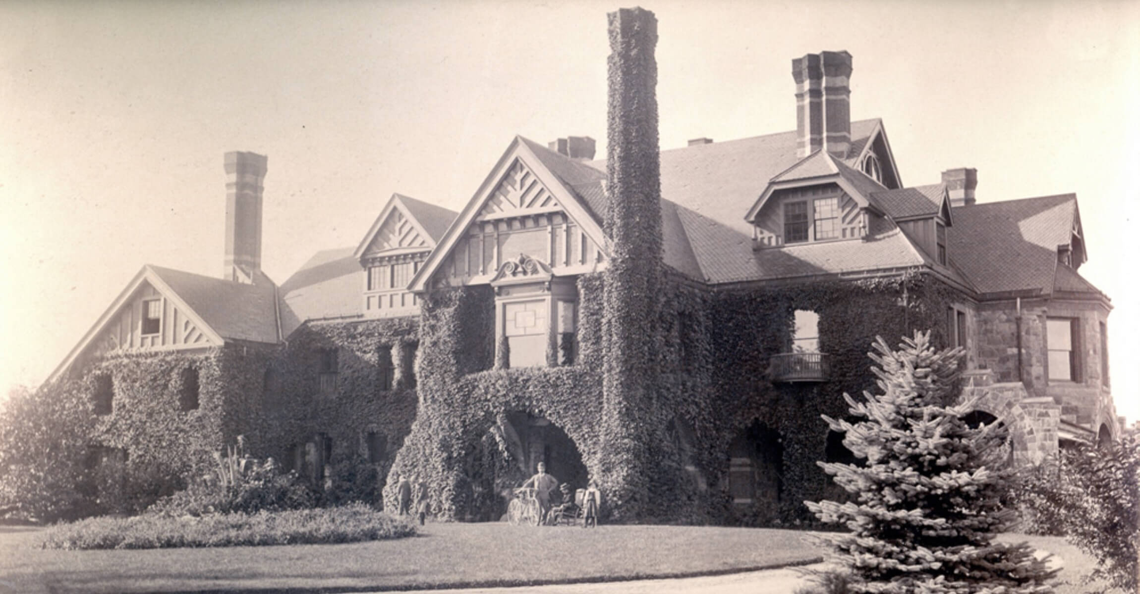 Black and white photograph of exterior of large stone house covered in ivy. In front of house are a man, woman, young girl and two teenage boys.