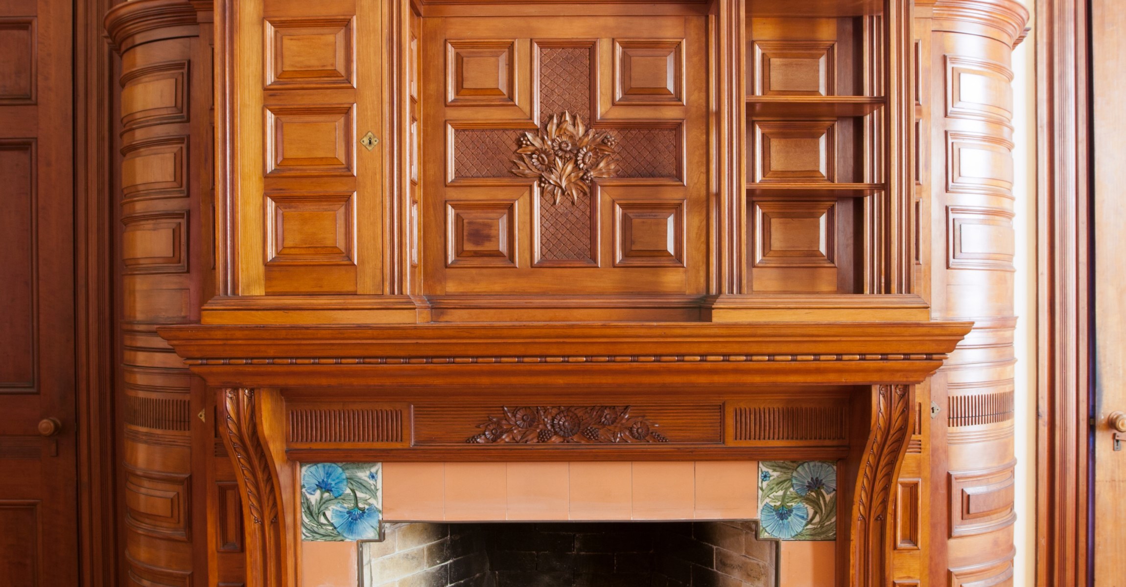 Wood paneled fireplace mantel with carved floral decorations and blue flower tiles.