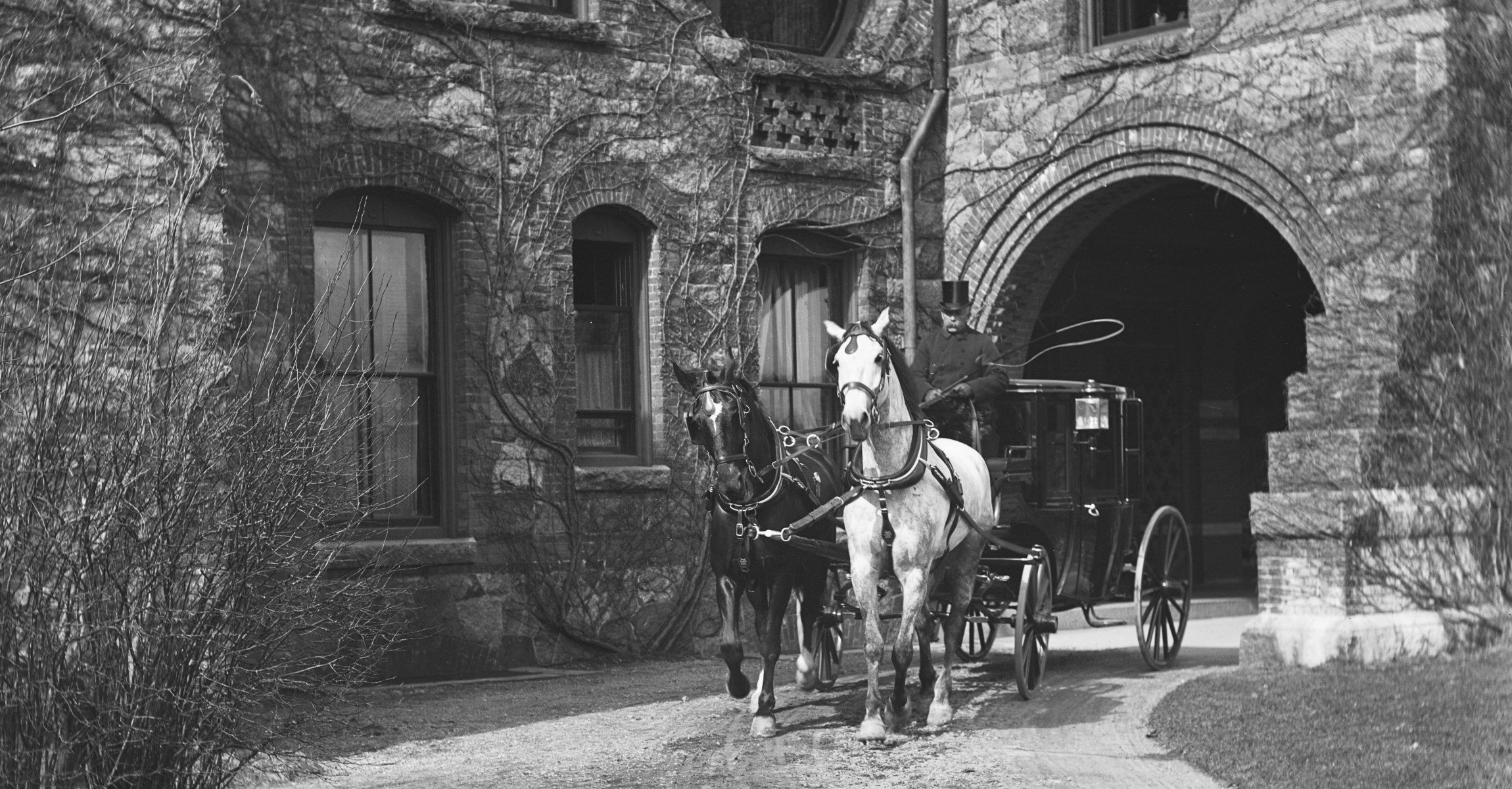 Black and white photograph of a carriage pulled by one black and one white horse with a driver wearing a top hat in front of stone house near archway.