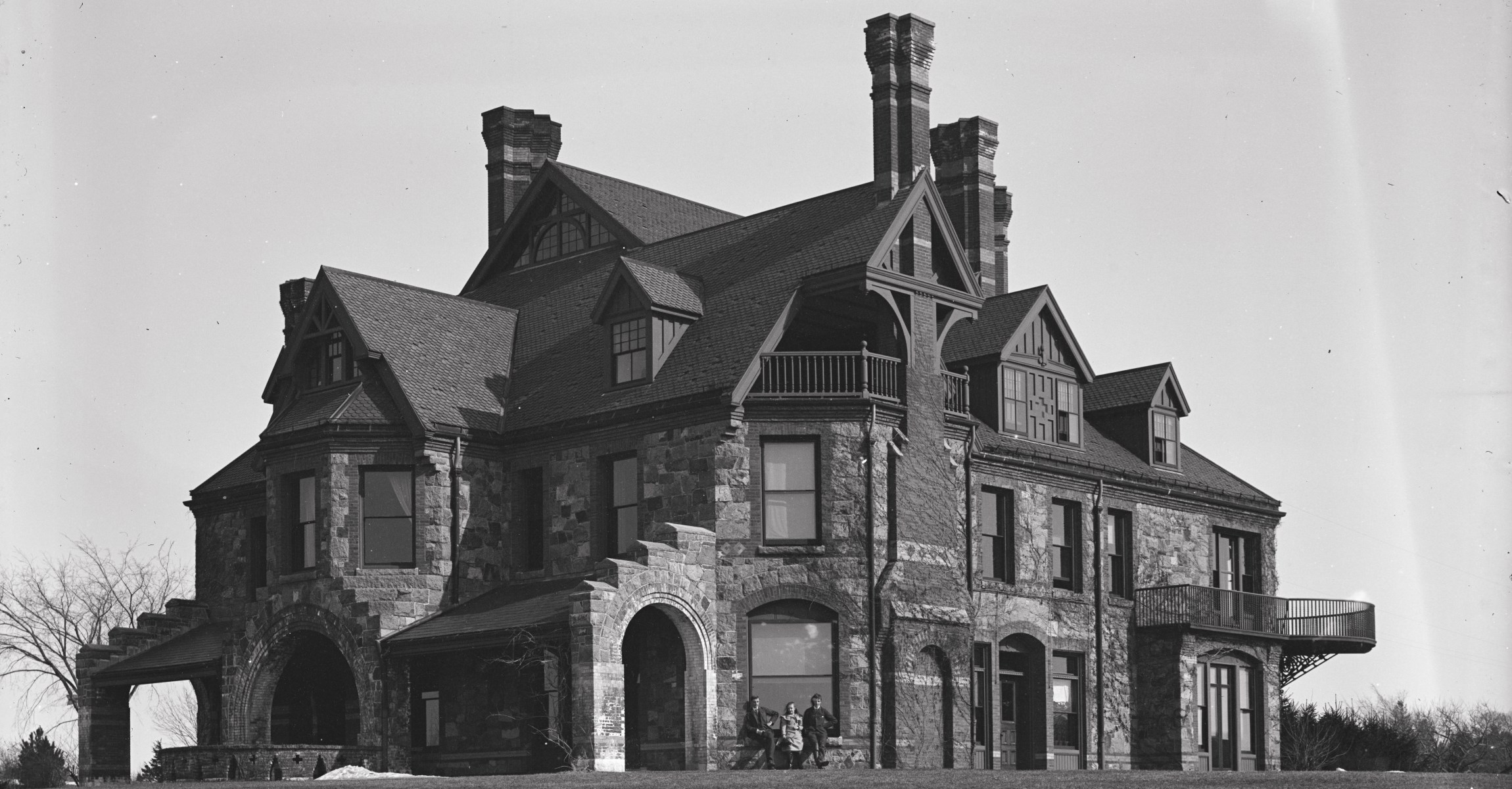 Black and white photograph of large stone house with many chimneys and balconies. Two teenage boys and a young girl pose in front of a window and appear very small.