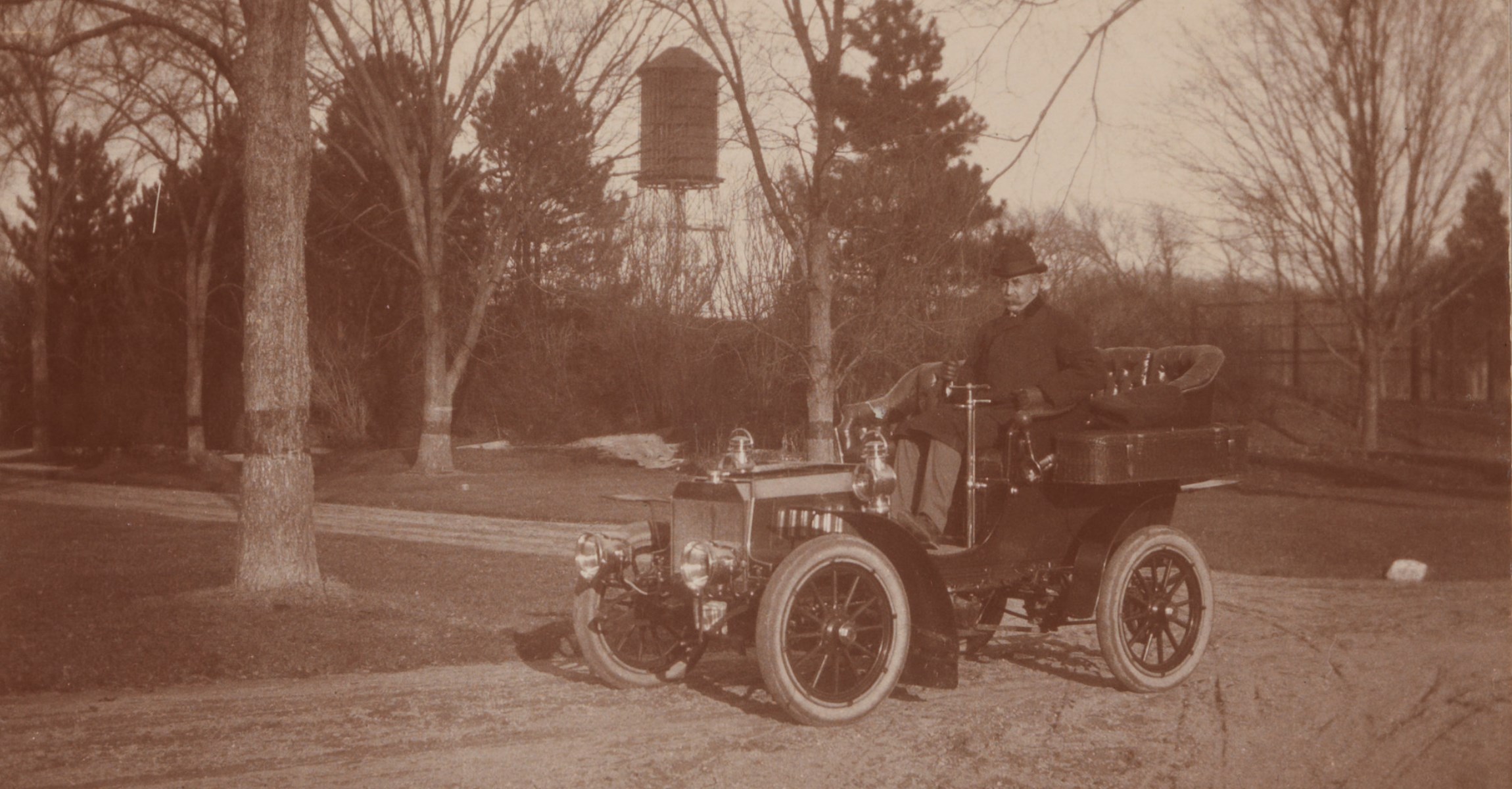 Sepia photograph of man with moustache and bowler hat in open top automobile. Wind turbine is visible in distance.