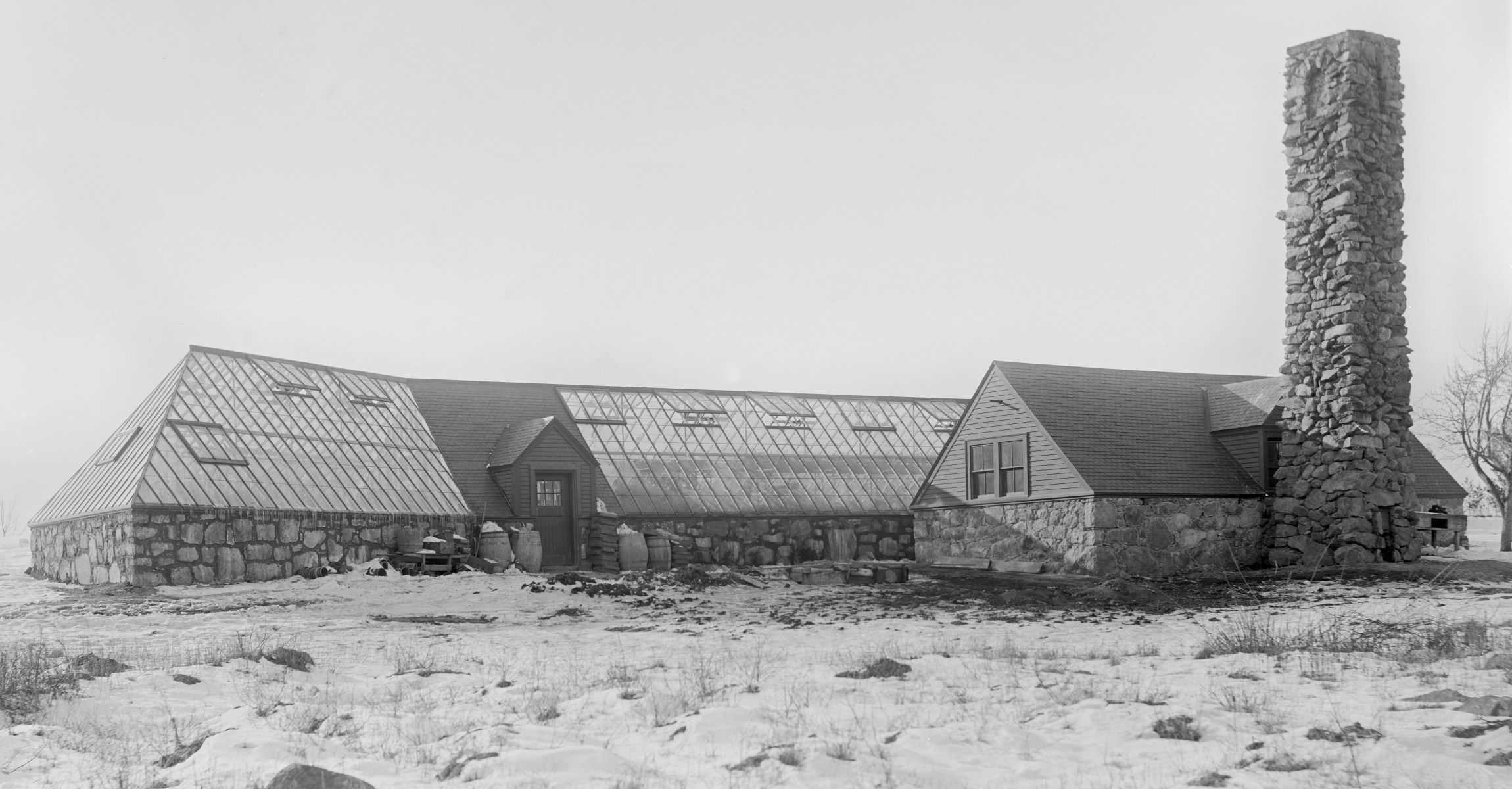 Black and white photograph of greenhouse with large stones along bottom of building and slanting glass panel roof.