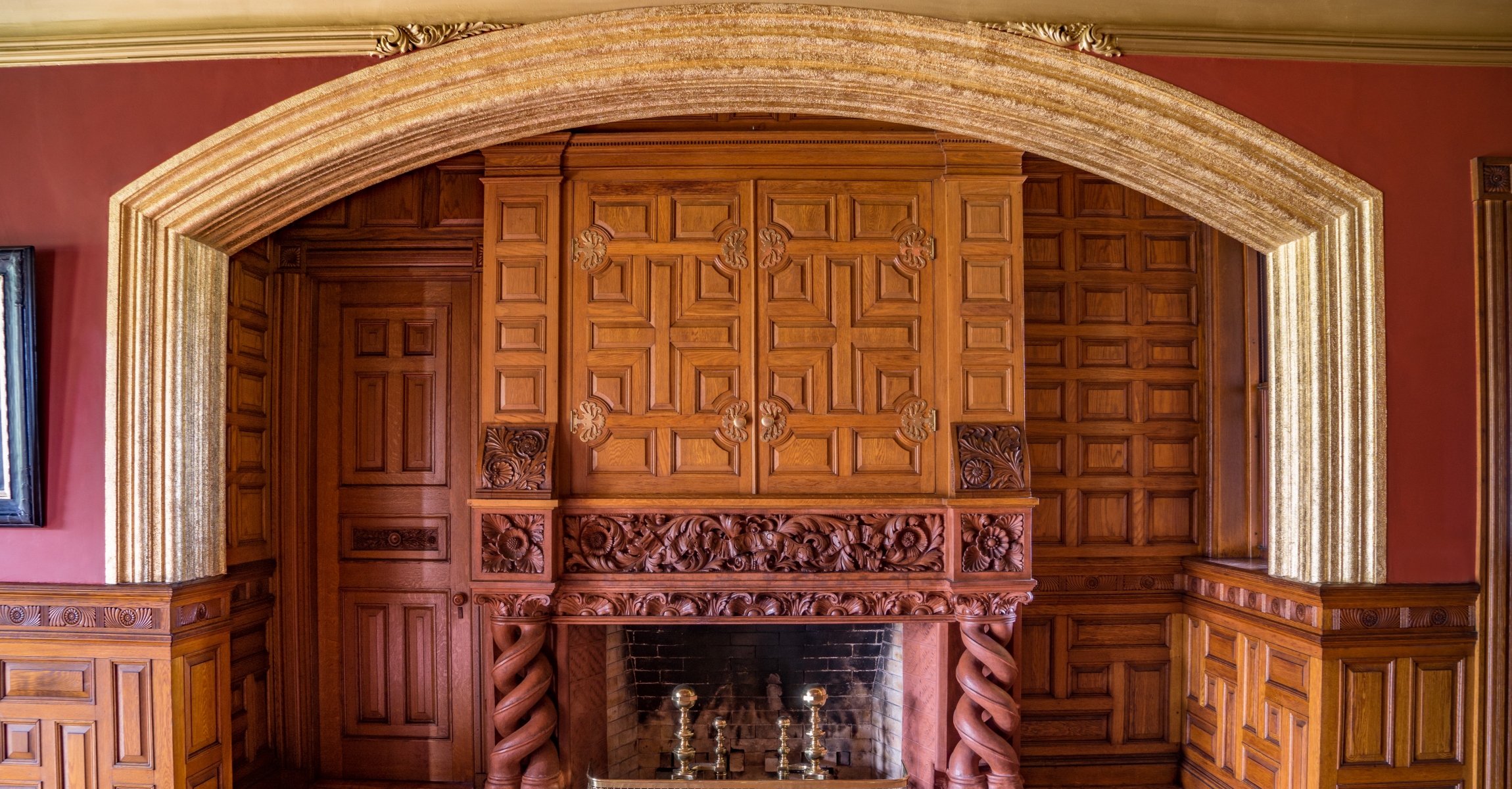 Large fireplace set into niche of a wood paneled room with brick red walls. There is a door to the left of the fireplace.