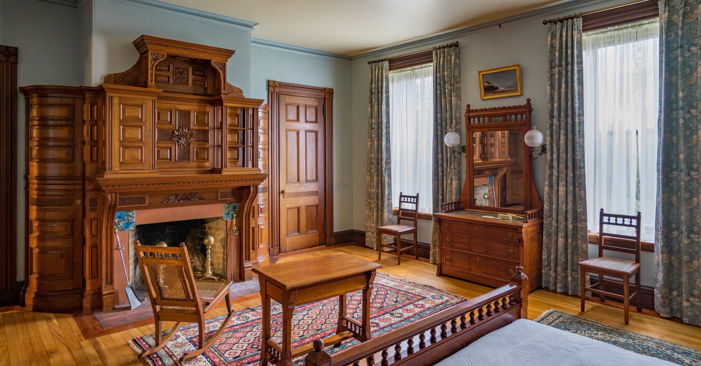 Room with wood fireplace mantle, wood bed, table, chairs, and dresser, patterned carpet and curtains, and blue walls with a painting hanging above mirror. 