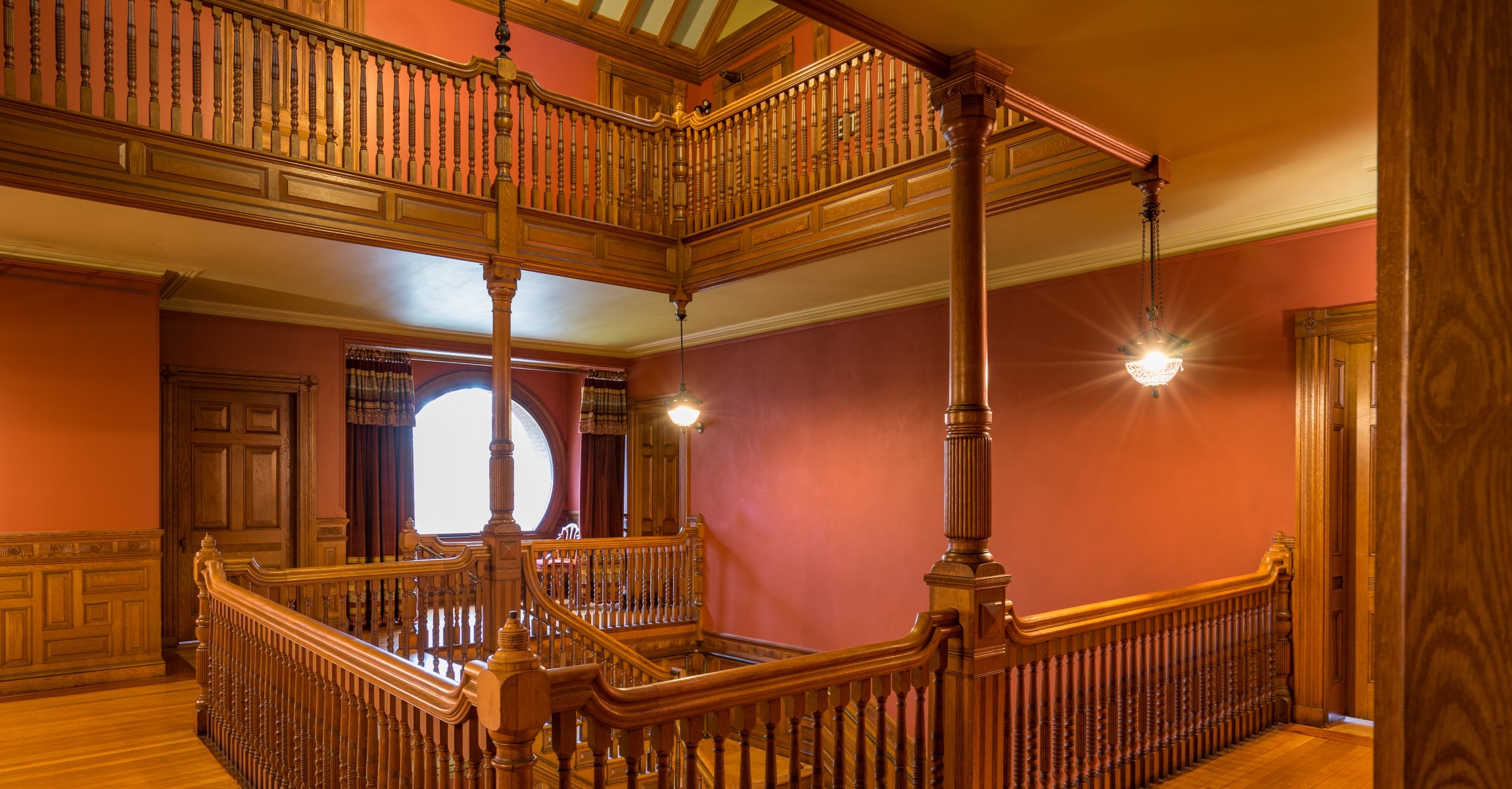 Wood railings around top of staircase and around the floor above, wood columns, hanging light fixtures, round window surrounded by maroon curtains.