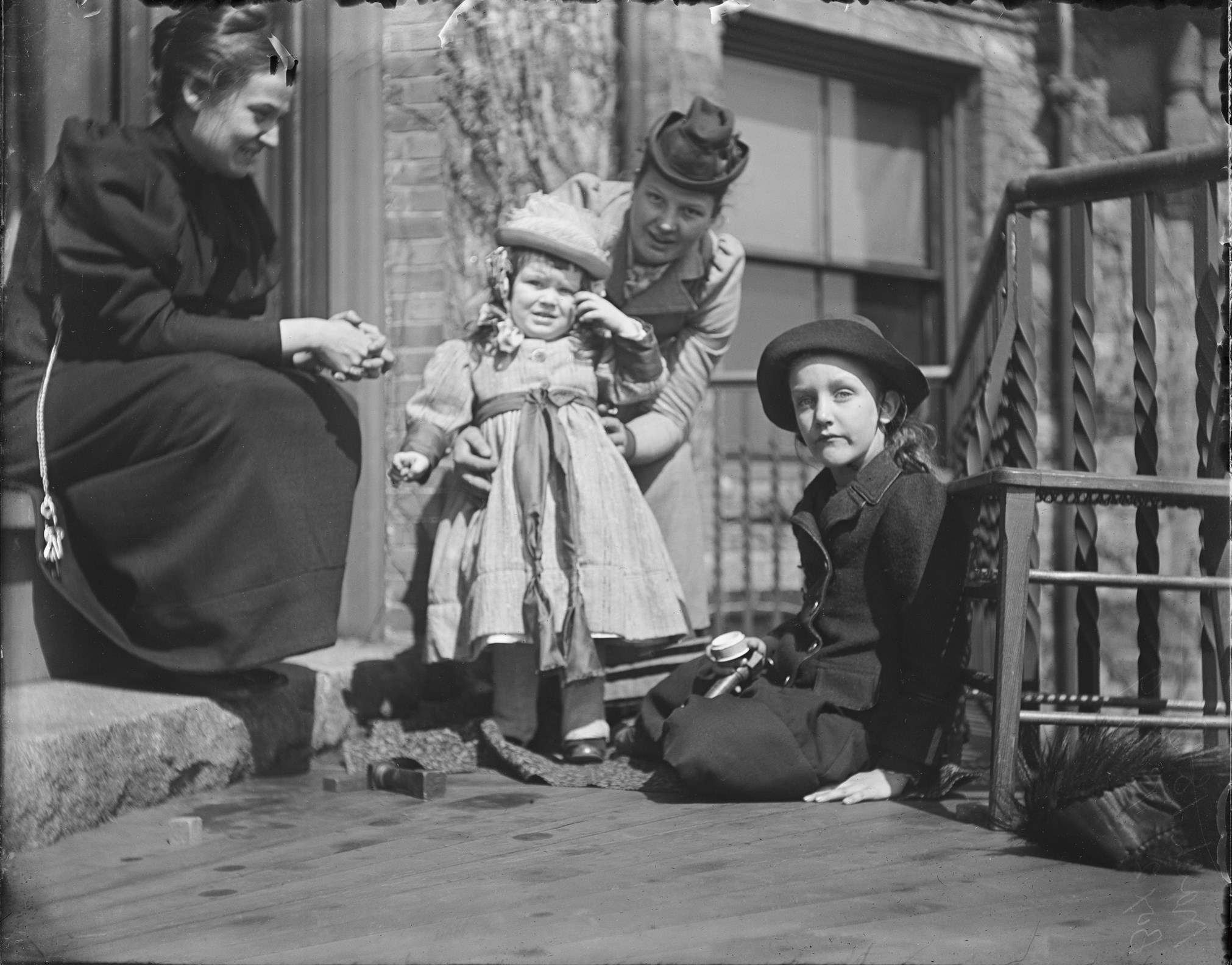 Black and white photograph of seated woman in black dress, standing woman in lighter colored coat and hat, small child in dress and hat, and young girl in dark coat and hat sitting on the balcony.