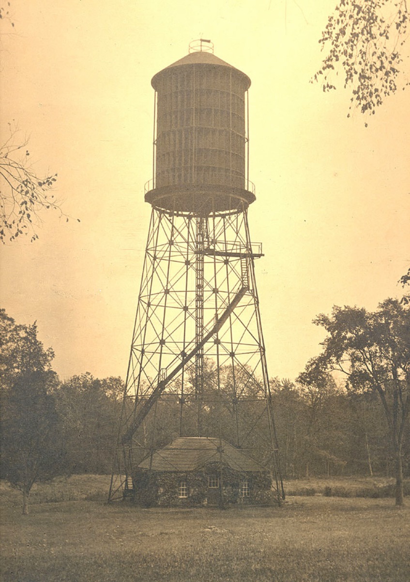 Sepia photograph of cylindrical windmill on tall steel scaffolding with ladders curving up the side, positioned directly over a one story stone building.