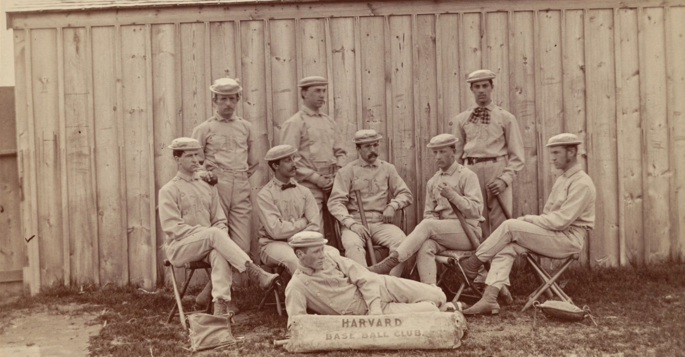 Black and white photograph of nine men in baseball uniforms, some hold bats and balls. One man laying on ground behind sign reading Harvard Base Ball Club.