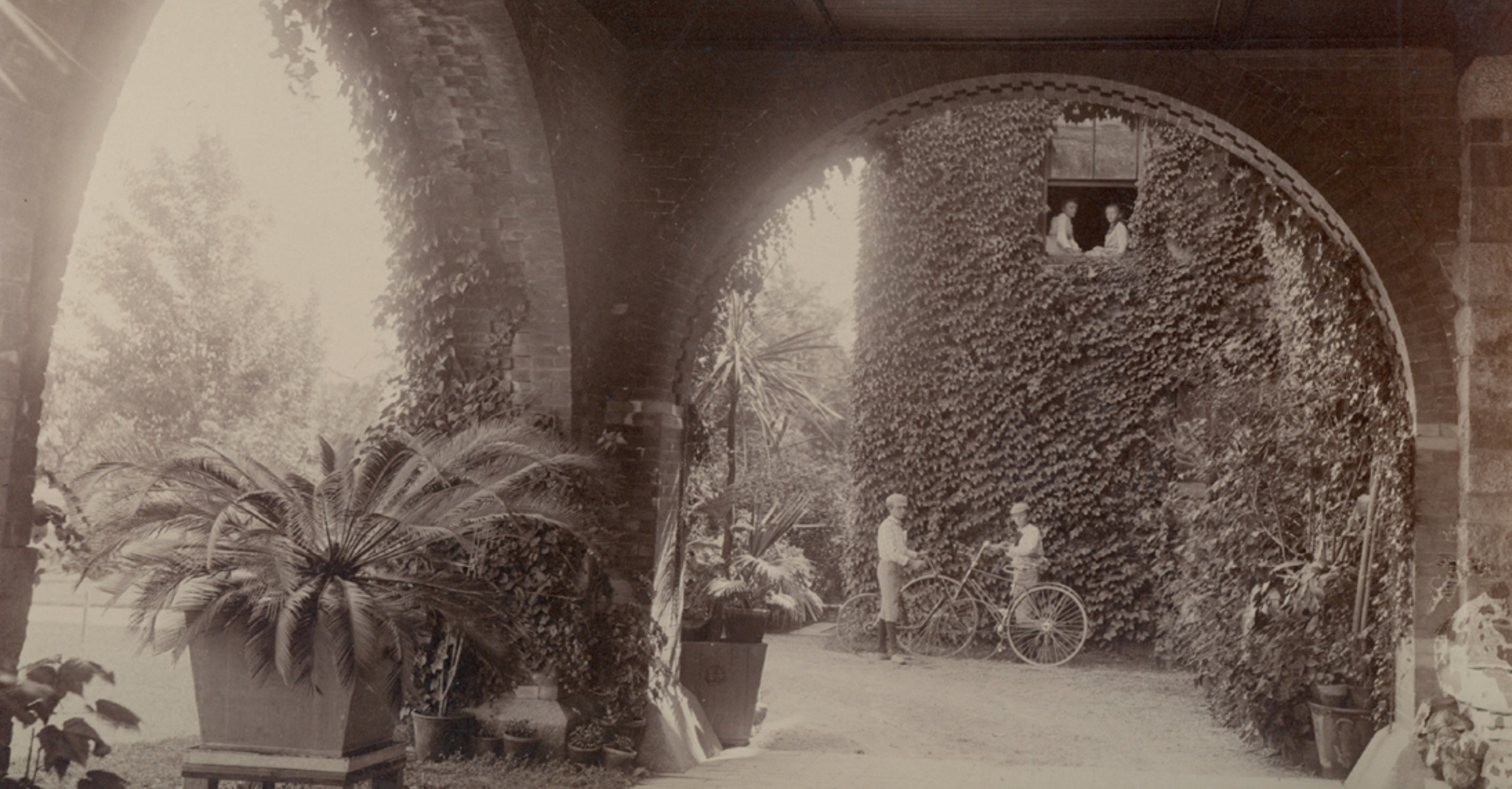 Black and white photograph looking through archway at ivy covered wall with two young boys with bicycles and two girls looking out an open window above them.