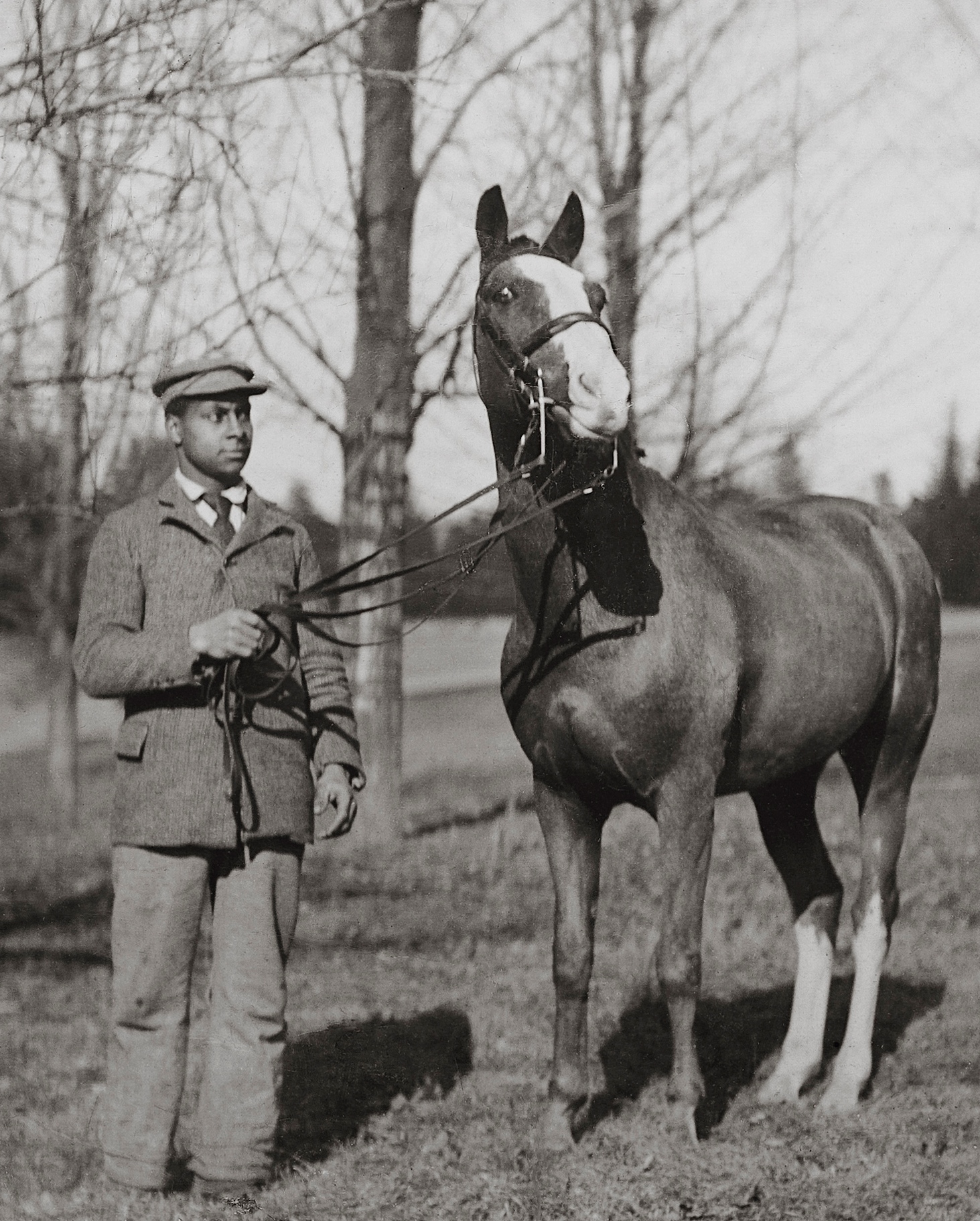 Black and white photograph of an African American man in his twenties in coat, tie, and hat, holding the reins of a horse outside.