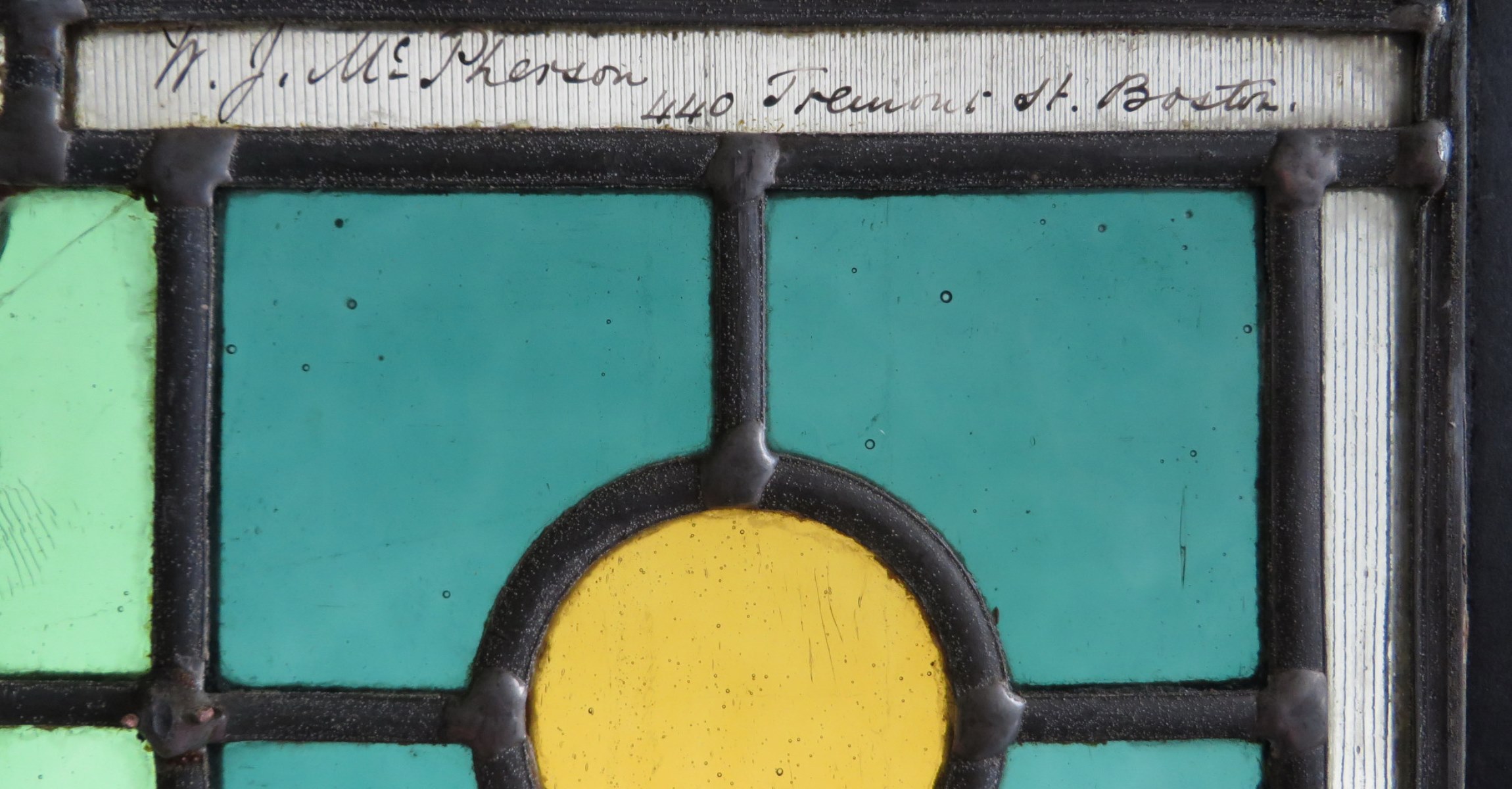 Close up view of stained glass window with black leading dividing panes of green, blue, and yellow glass. Writing along top of the panel.