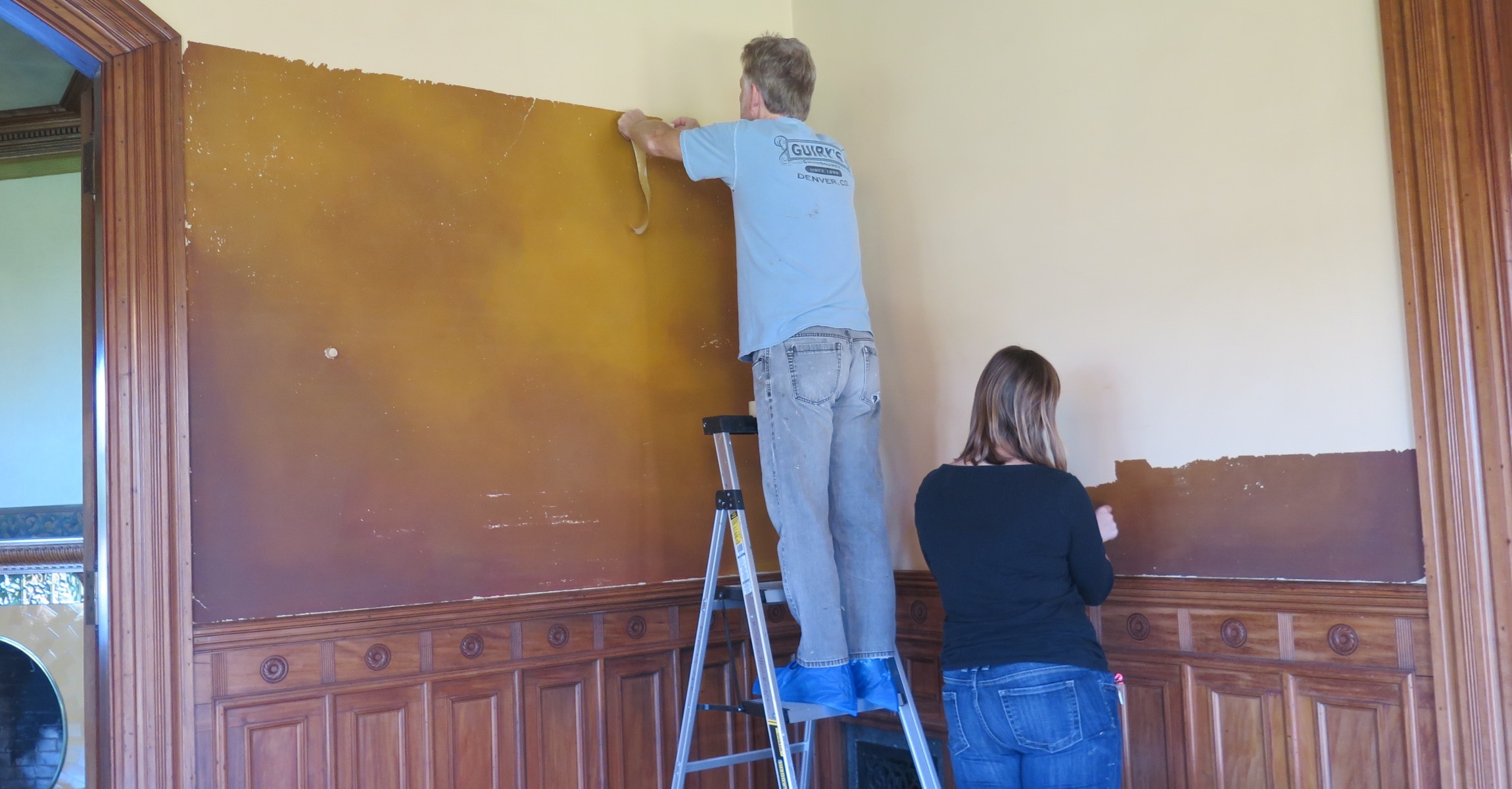 A man on a ladder and a woman on the ground facing a wall that is half white and half amber.