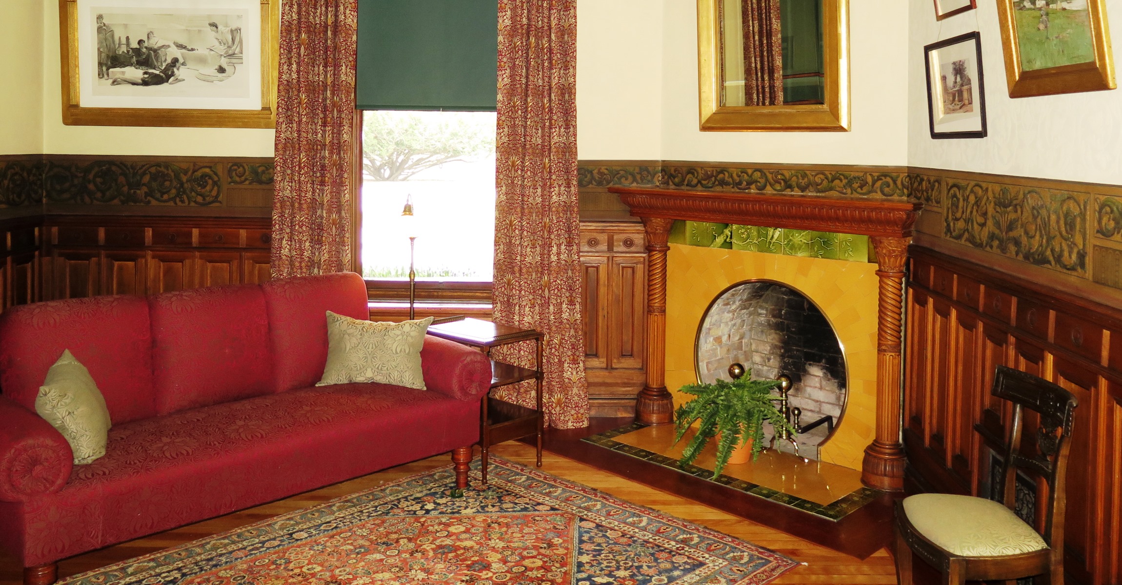 Room with a red sofa, patterned carpet and curtains, wood paneling, paintings on the wall, decorative paint border, and fireplace with round opening and green and yellow decorative tiles.