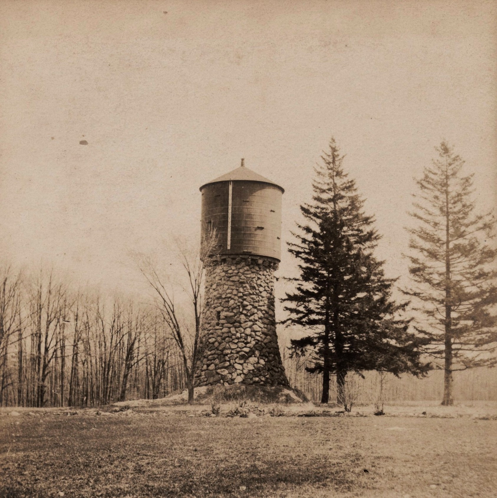 Black and white photograph of a cylindrical water tower atop a tall stone base next pine trees.