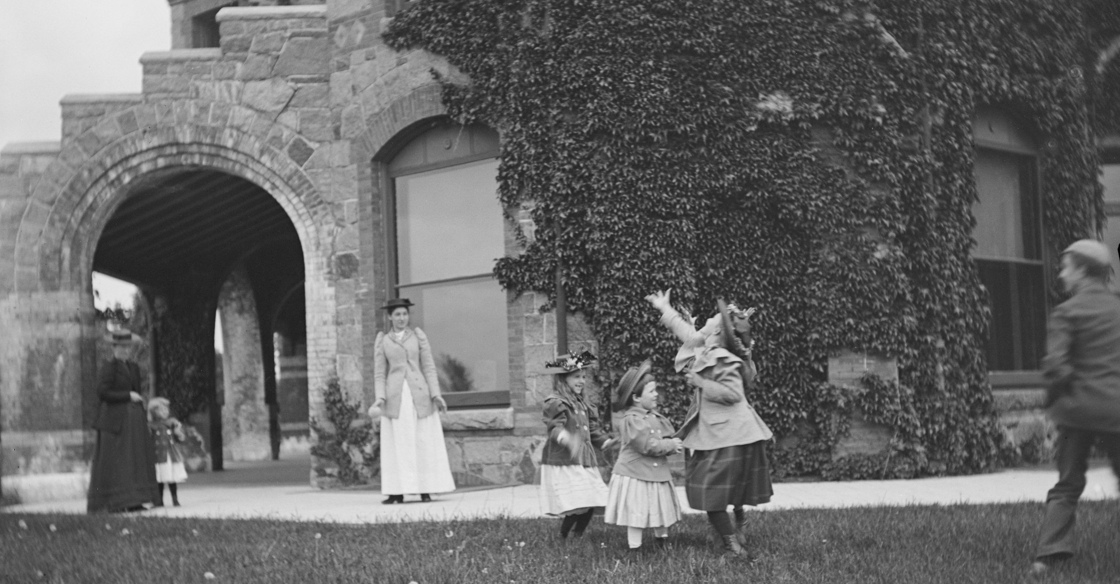 Black and white photograph of children playing outside stone building covered with ivy.