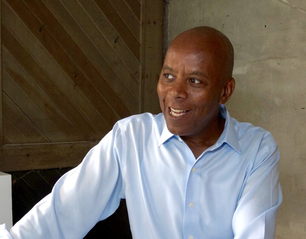 Photograph of artist Richard Haynes, a smiling Black man in a light blue collared shirt in front of a beige wall.