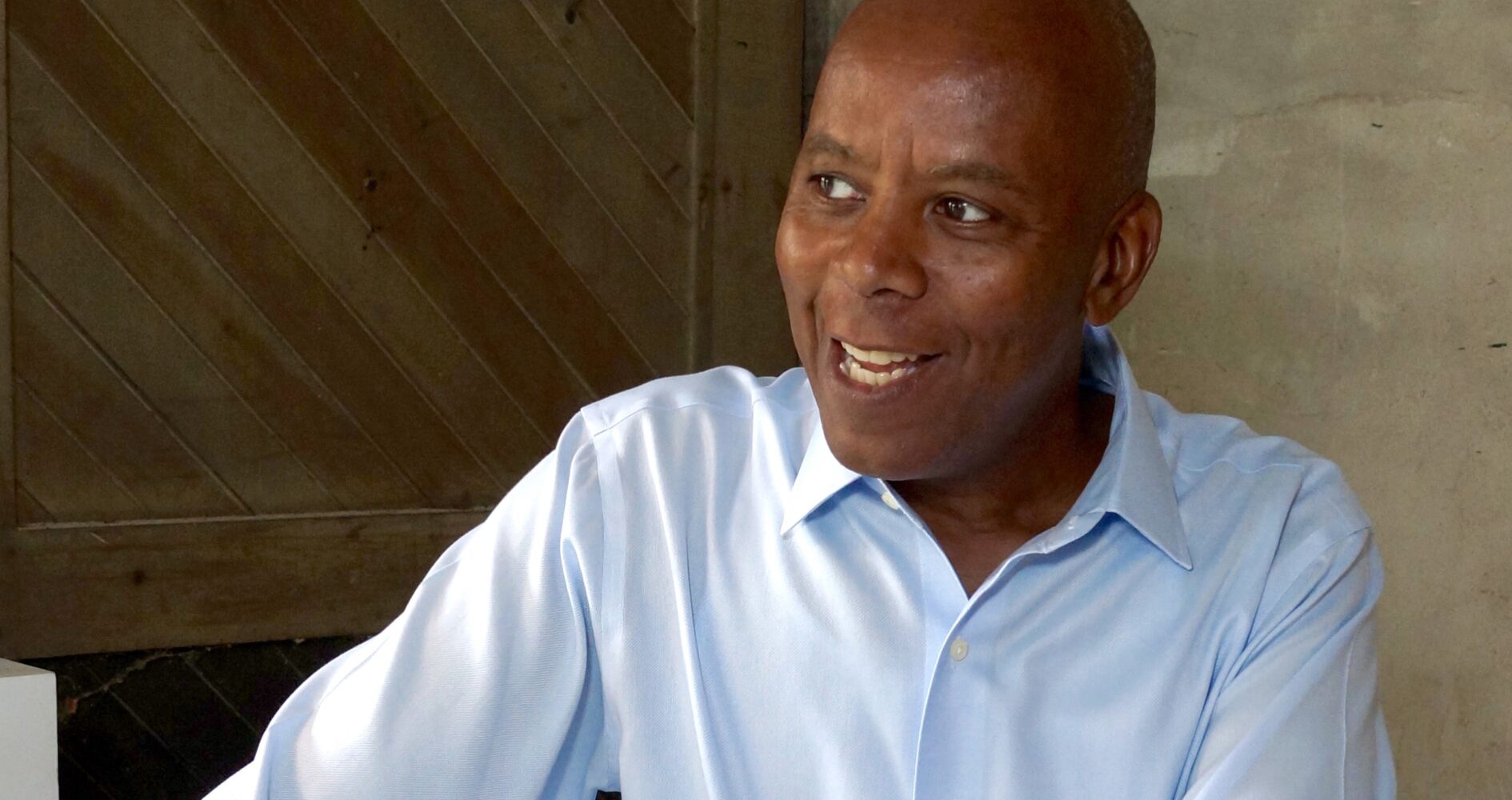 Photograph of artist Richard Haynes, a smiling Black man in a light blue collared shirt in front of a beige wall.