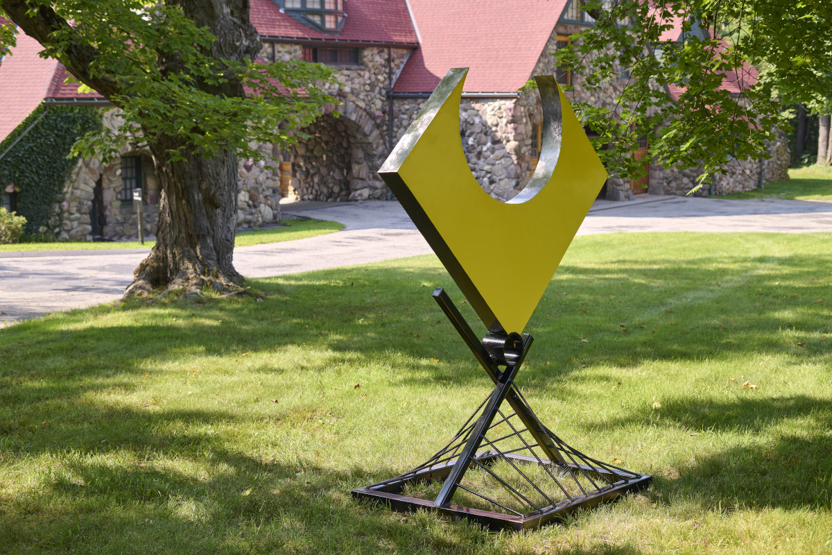 Yellow and black geometric sculpture on grass in front of a stone building and trees.