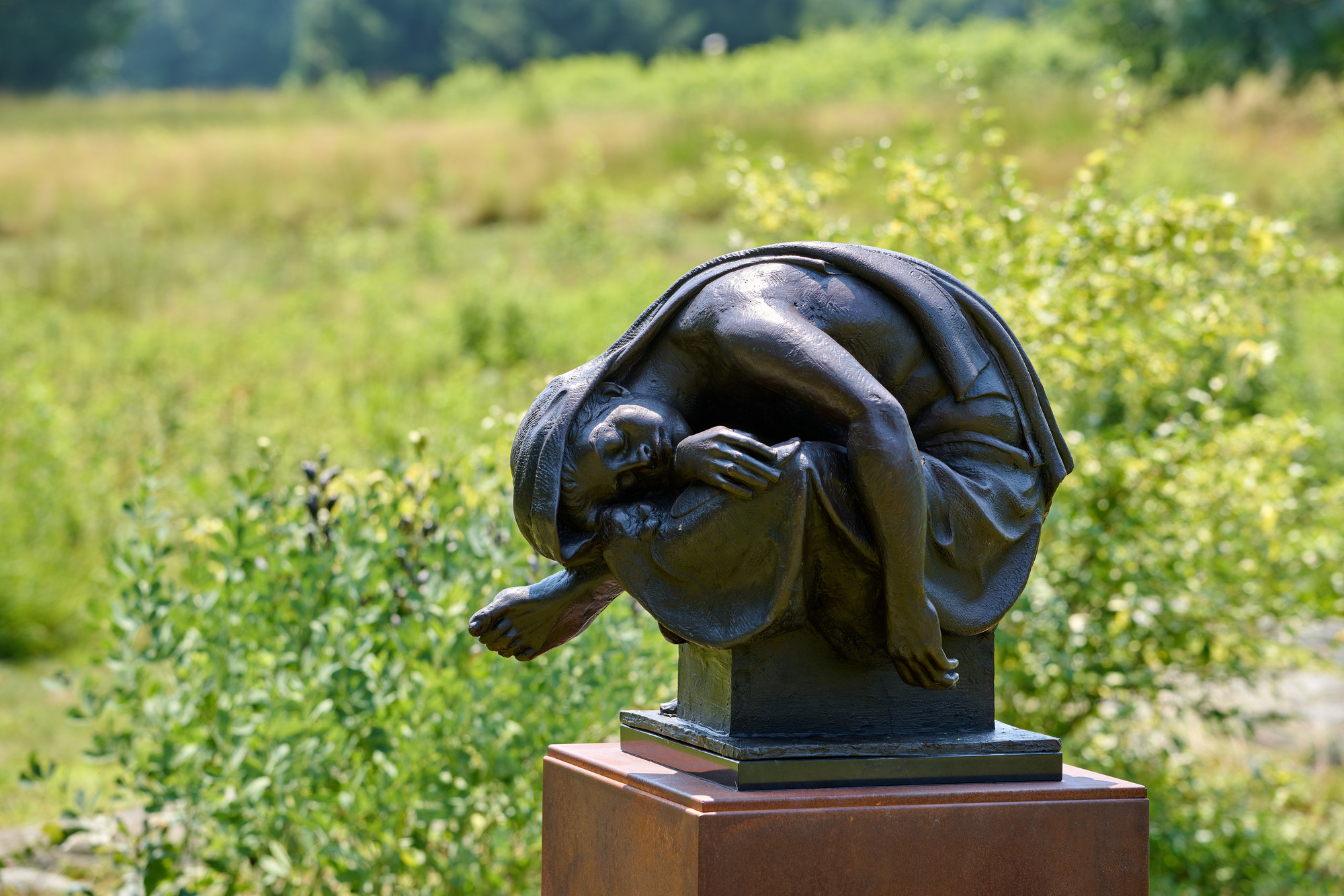 Bronze sculpture of a sleeping woman of color doubled over and resting on her leg.  Sculpture is outside with green meadow in the background.