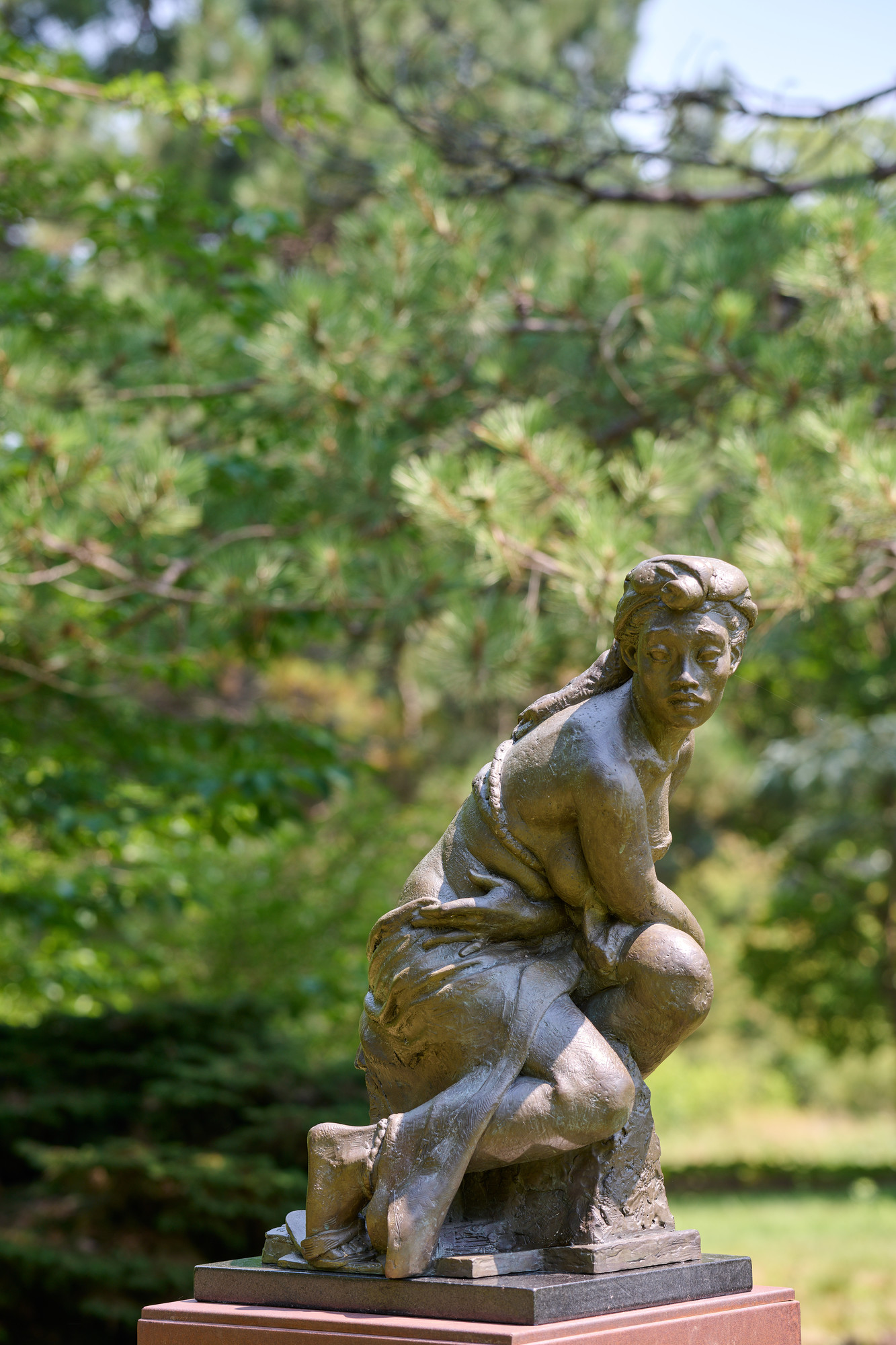 Bronze sculpture of a woman of color draped in fabric and seated on a rock, turning to look over her shoulder. Sculpture is outside with pine trees in background.