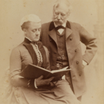 Man with mustache standing and looking over shoulder of woman with white cap holding a book.